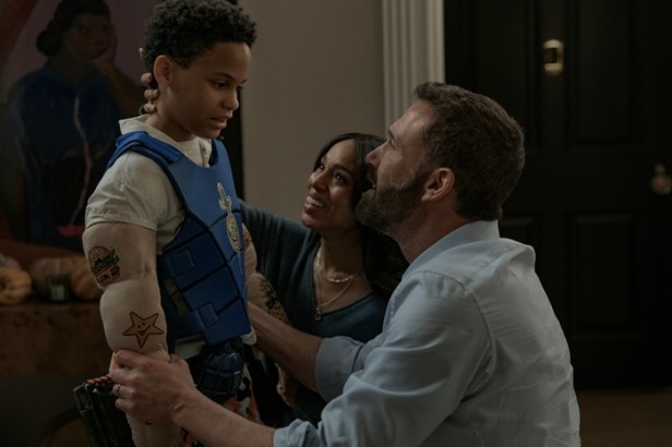 "A child wearing a muscle costume and blue vest stands while smiling adults, a man kneeling and a woman crouching, look at him warmly in a cozy, dimly lit room with artwork and furniture in the background."
